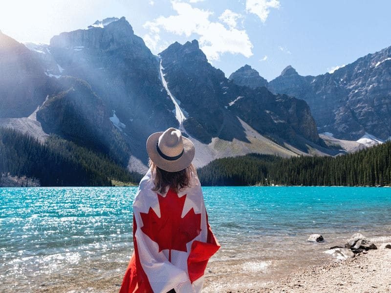 Woman wrapped in Canadian flag looks across lake at mountains.